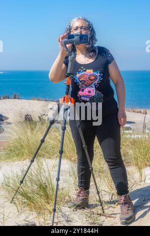 Femme mature concentrée programmant son téléphone portable avec stabilisateur de tête de trépied pour prendre un film, des dunes, la plage et la mer en arrière-plan, journée ensoleillée dedans Banque D'Images