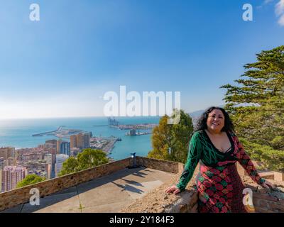 Femme d'âge moyen posant sur la terrasse du point de vue à Alcazaba de Malaga, expression souriante, longs cheveux noirs, portant une robe multicolore, paysage urbain, seap Banque D'Images