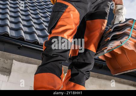 Un ouvrier de la construction en tenue orange et noire équilibre sur un toit, portant des tuiles de toit pour l'installation pendant une journée brillante sur un chantier de construction. Banque D'Images