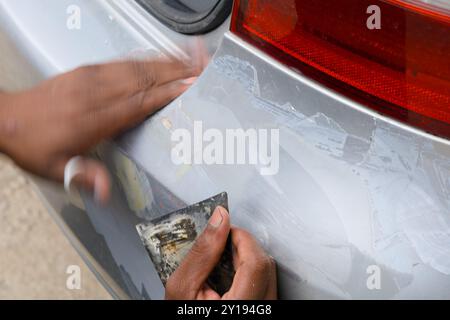 Une personne applique méticuleusement du mastic pour lisser les rayures sur un pare-chocs de voiture dans un garage bien éclairé. Outils et matériels sont organisés à proximité, signif Banque D'Images