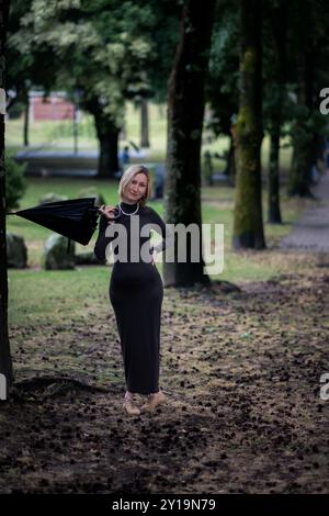 Une femme tenant un parapluie plié se tient près de grands pins dans un parc de la ville. Banque D'Images