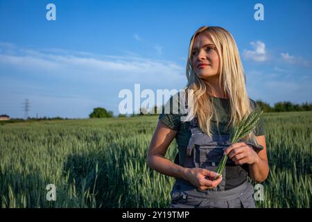 Agricultrice satisfaite et souriante debout dans un champ de céréales, tenant des tiges de blé dans ses mains Banque D'Images