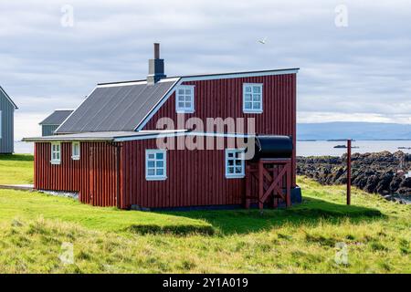 Maison peinte en rouge, montagnes en arrière-plan et l'océan contre ciel nuageux. Flatey, Breiðafjörður, Westfjords, Islande Banque D'Images