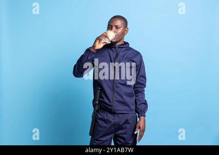 Portrait de l'agent de sécurité afro-américain boit une tasse de café en studio, profitant d'un rafraîchissement de caféine sur fond bleu. Employé armé confiant servant une boisson. Banque D'Images