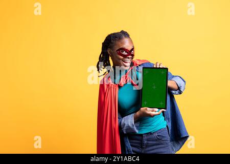 Fille portant le costume de super-héros et le masque pour Halloween mettant en vedette la tablette de clé chroma, fond de studio. Portrait d'une femme habillée comme héros de bande dessinée cachant l'identité présentant un dispositif d'écran vert Banque D'Images