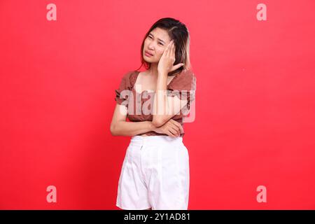 Expression d'une femme indonésienne tenant sa tête dans la douleur, franche, portant une blouse brune et un short avec un fond rouge. pour la santé, les soins et l'adver Banque D'Images