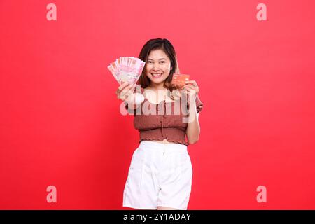 Joyeuse fille asiatique tenant de l'argent rupiah devant elle et une carte de crédit de débit portant une blouse brune avec un fond rouge. pour la transaction, techno Banque D'Images