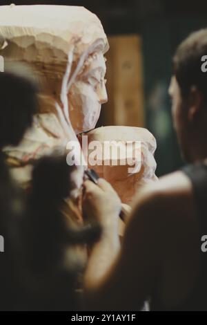 Un artisan travaille sur une sculpture en bois détaillée d'une femme et d'un enfant dans un atelier, capturée de profil Banque D'Images