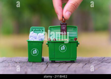 Gros plan d'une main plaçant une bouteille en verre dans un bac de recyclage vert miniature, soulignant l'importance du recyclage et de l'élimination responsable des déchets. Banque D'Images