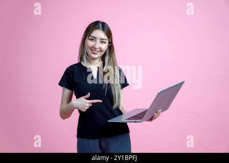 Jeune femme asiatique debout avec un sourire, pointant vers le moniteur d'ordinateur portable. Portrait sur fond rose avec lumière de studio. Banque D'Images