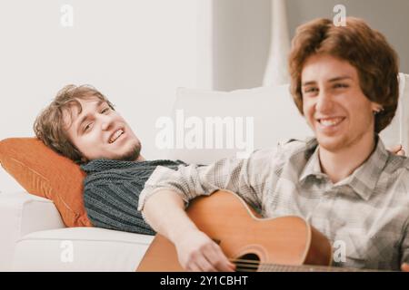 Deux amis se prélassant sur un canapé, jouant de la guitare, riant et appréciant le confort de la maison. Partager musique et joie dans un salon moderne. Temps de qualité avec Banque D'Images