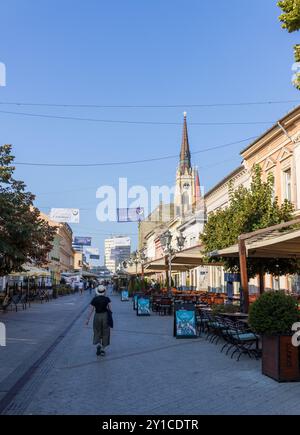 Une femme touriste dans un chapeau marchant à côté de cafés et restaurants sur la rue Zmaj Jovina dans la ville serbe de Novi Sad par une journée ensoleillée avec un ciel bleu. Banque D'Images