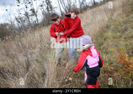 Deux garçons en vestes rouges s'engagent dans la lutte ludique dans un champ herbeux tandis qu'une jeune fille en tenue d'hiver rigole et les regarde. L'atmosphère est Banque D'Images