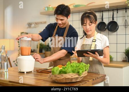 Heureux jeune couple préparant des smoothies sains et suivant une recette en ligne sur tablette numérique Banque D'Images