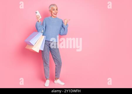 Photo pleine longueur des sacs à provisions de dispositif d'espace vide de point de look féminin à la retraite portent une tenue en tricot bleu isolée sur fond de couleur rose Banque D'Images