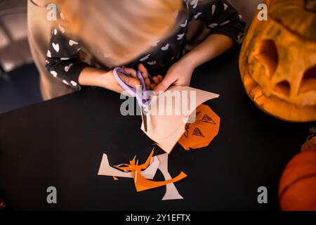 Un enfant découpe diligemment des formes d'Halloween dans du papier orange et beige à une table. A proximité, une citrouille sculptée ajoute à l'ambiance festive, tandis que co Banque D'Images
