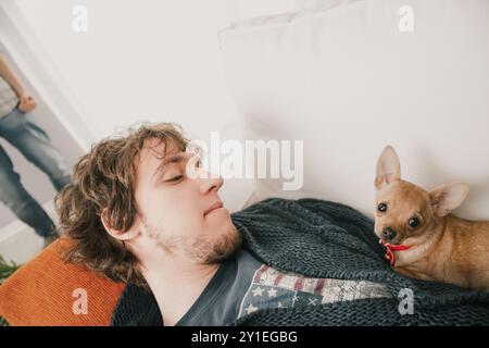 Le jeune homme aux cheveux longs est allongé sur un canapé les yeux ouverts, regardant loin de la caméra tandis qu'un petit chien chihuahua brun repose la tête sur ses CHES Banque D'Images