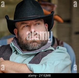 Homme blanc caucasien avec barbe et chapeau de cow-boy noir est assis et semble réfléchi et grincheux. Il rince en levant les yeux et en s'éloignant sur le côté, les bras pliés. Banque D'Images