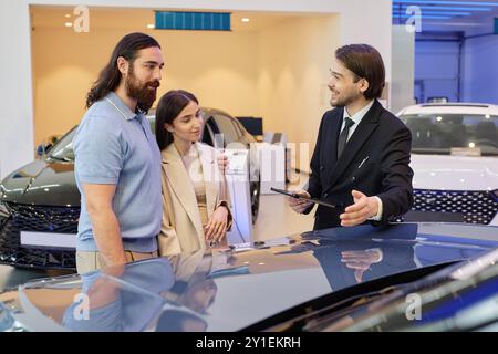 Plan moyen d'un concessionnaire automobile souriant d'Asie centrale montrant un homme adulte et une femme nouveau véhicule tout en conseillant couple sur l'achat d'une nouvelle voiture au centre de concession Banque D'Images