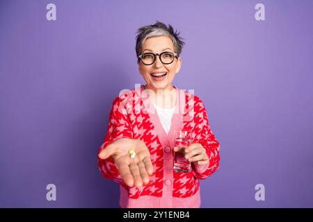 Portrait photo de belle dame retraitée tenir des pilules pharmacie boire de l'eau habillée élégant vêtement rouge isolé sur fond de couleur violette Banque D'Images