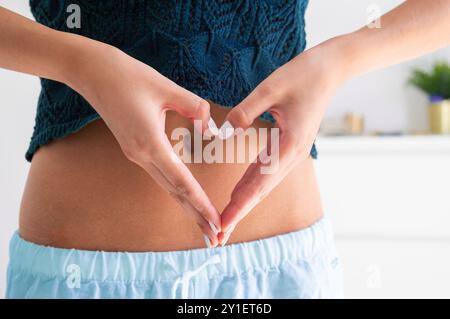 détail d'une jeune femme habillée décontractée dans un haut tricoté et un pantalon en lin fait une forme de coeur avec ses mains devant son ventre plat. Les soins de santé Banque D'Images