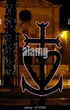 Aigues-mortes, Frankreich, 25,12 .2023 Das Wahrzeichen der Camarque Croix camarguaise setzt sich aus den Symbolen für Glaube,Liebe und Hoffnung zusammen und ist in der Weihnachtszeit auf dem Vorplatz der Chapelle de la confrÃ rie des PÃ nitents-gris beleuchtet aufgestellt. Es verkörpert die drei theologischen Tugenden : Glaube, Hoffnung und Liebe. Es symbolisiert die Camargue, weil das Kreuz mit den Dreizacken Glauben an die Gardian mit ihrem Dreizack-Spieß erinnert, der Anker Hoffnung an die Fischer der Camargue und das Herz Liebe an die heiligen Marien Maria Magdalena, Maria Kleophae und Maria Banque D'Images