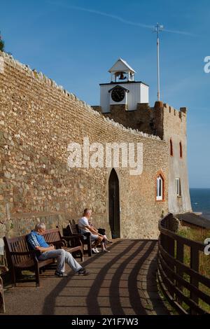 Les gens assis sur des bancs sur le chemin sous la Tour de l'horloge et Flint Wall de Connaught Gardens, Sidmouth, Royaume-Uni Banque D'Images