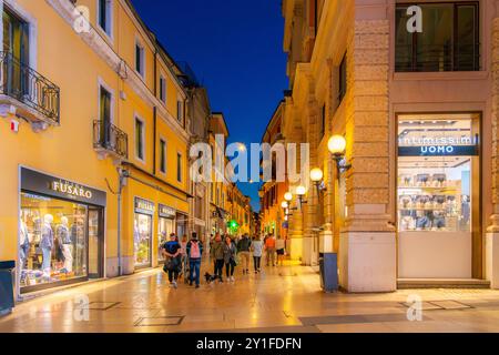 Les piétons parcourent et visitent la rue illuminée via Giuseppe Mazzini, qui regorge de boutiques, de cafés et de boutiques dans la vieille ville historique de Vérone, en Italie. Banque D'Images