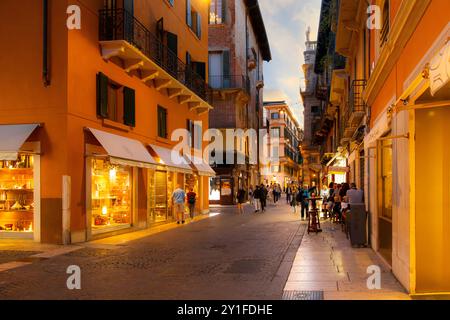 Les piétons parcourent et visitent une rue illuminée de magasins, boutiques et cafés dans le centre historique de la vieille ville de Vérone, en Italie Banque D'Images