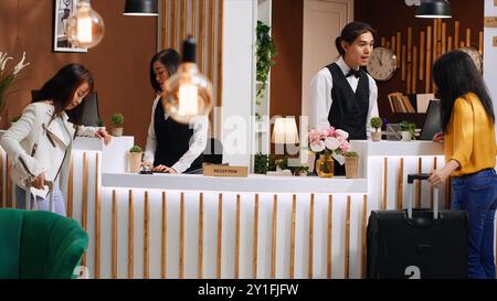 Jeune client approchant la réception dans le hall de l'hôtel, se préparant à l'enregistrement après un long vol. Le personnel asiatique accueille les clients à la réception, discutant de la réservation de chambre et des installations. Banque D'Images