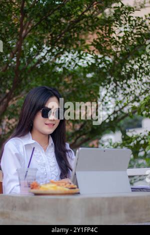 bureau belle femme asiatique dans un café en plein air. fille souriante faisant le travail sur ordinateur portable dans des lunettes de soleil candid pour le contenu ou la publicité Banque D'Images