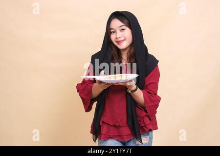 Belle femme asiatique mature portant un hijab rouge souriant tout en portant une assiette contenant dimsum (nourriture chinoise) et baguettes tout en servant. utilisé fo Banque D'Images