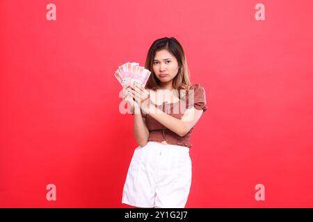 L'expression de la femme asiatique est sullen, les deux mains tenant la monnaie rupiah, portant une blouse brune, fond rouge. pour les transactions, la technologie et la publicité Banque D'Images