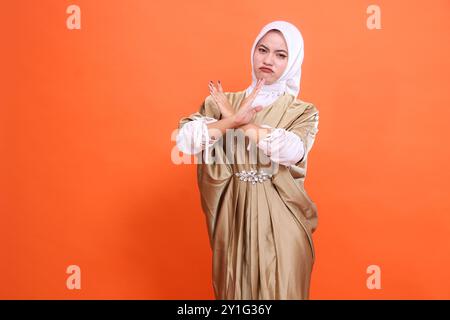 Expression d'une belle femme asiatique adulte frôlant les sourcils avec les bras croisés devant un panneau stop portant une robe musulmane caftan avec un hijab, isolez Banque D'Images