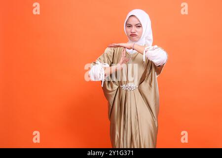 expression de la belle femme adulte d'indonésie froncant les sourcils signe d'arrêt portant robe musulmane de caftan avec hijab, isolé sur fond orange. Concept Banque D'Images