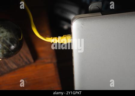 Vue rapprochée d'un câble Ethernet jaune branché sur un ordinateur portable argenté. Le fond comporte une surface en bois et un objet décoratif, ajoutant un co Banque D'Images