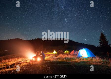Camping de nuit dans les montagnes sous le ciel étoilé. Silhouette de deux personnes, couple assis sur l'herbe près du camping, admirant le paysage. Concept de tourisme et de vie saine. Banque D'Images