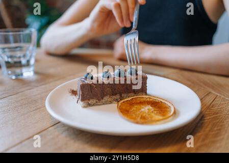 Femme se prépare à déguster un dessert au chocolat végétalien surmonté de myrtilles, accompagné d'une tranche d'orange séchée sur une assiette blanche avec fourchette dans un café sur une table en bois Banque D'Images