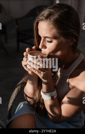 Une femme aux longs cheveux bruns est assise sur une chaise et boit dans une tasse blanche. La lumière du soleil brille sur son visage et son bras. Elle porte un débardeur blanc et une tanière bleue Banque D'Images