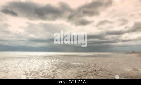 Une plage de sable à marée basse avec une mer chatoyante qui s'étend à l'horizon. Un grand troupeau d'oiseaux sont sur la mer et des nuages sombres dramatiques sont au-dessus. Banque D'Images