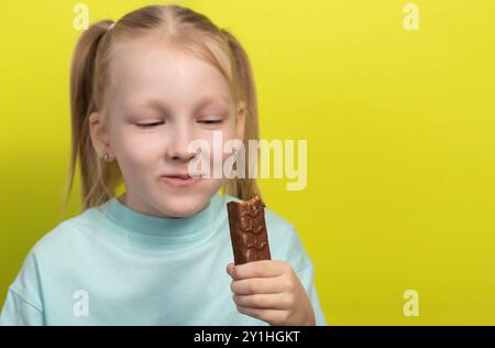 Une belle fille joyeuse aux cheveux blancs de sept ans mange une barre de chocolat sur fond jaune. Les enfants sont des amoureux de la malbouffe sucrée, horizonto Banque D'Images