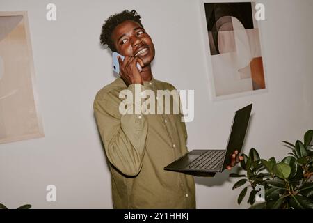 Jeune homme d'affaires tenant un ordinateur portable tout en parlant au téléphone avec des partenaires dans un bureau confortable Banque D'Images