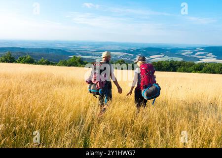 Deux randonneurs avec sacs à dos trekking, un couple marchant à travers une prairie herbeuse dorée vers une vue lointaine de collines ondulantes et un ciel bleu clair Banque D'Images