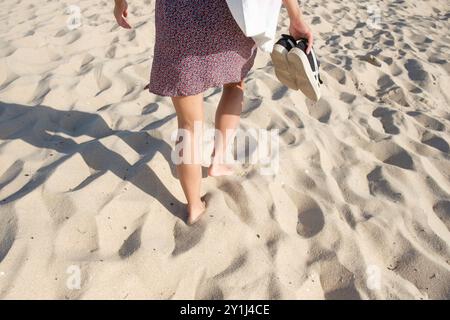 Femme en robe de soleil vue hanche vers le bas par derrière alors qu'elle marche pieds nus sur une plage de sable, tenant ses baskets dans une main. Banque D'Images