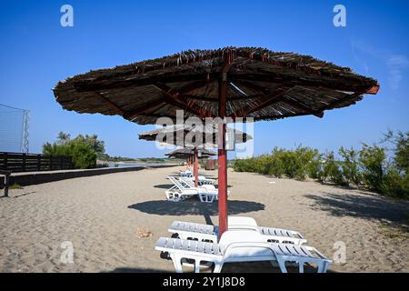 Chaises longues en plastique blanc debout sur une plage de sable avec des parasols de paille par une journée ensoleillée claire. Banque D'Images