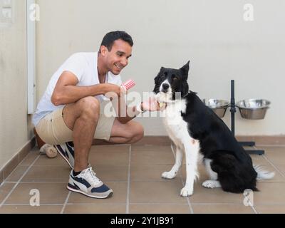 Un homme caresse un chien tout en tenant un bâton de glace rouge et blanc. Le chien est assis sur le sol et il apprécie l'attention Banque D'Images