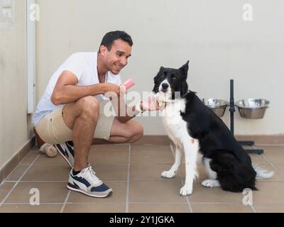 Un homme caresse un chien tout en tenant un bâton de glace rouge et blanc. Le chien est assis sur le sol et il apprécie l'attention Banque D'Images
