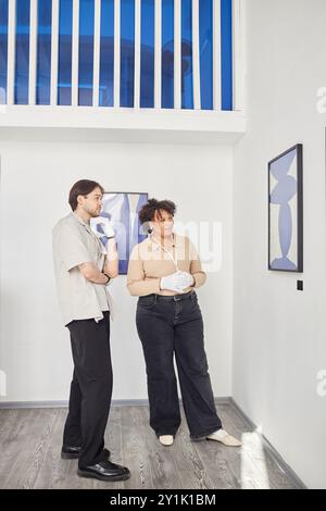 Portrait vertical pleine longueur de deux ouvriers de galerie d'art regardant des peintures d'art abstrait tout en planifiant une exposition dans le hall blanc du musée Banque D'Images