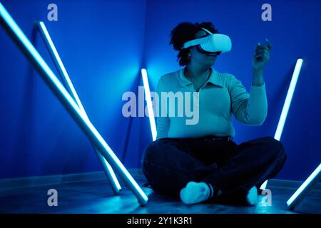 Portrait d'une femme portant un casque VR assis sur le sol dans des néons bleus et interagissant avec le cyberespace numérique Banque D'Images