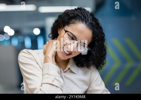 Femme d'affaires hispanique souffrant de douleurs au cou dans le bureau moderne. Elle semble stressée et mal à l'aise, probablement en raison d'un travail prolongé sur ordinateur. Concept de stress au travail et de tension physique. Banque D'Images